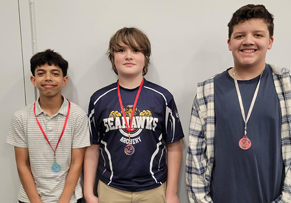 Three students standing side by side against a wall, each wearing a medal on a ribbon. The student in the center wears a “Seahawks Archery” shirt, and the others are dressed casually. All three face the camera, smiling or with neutral expressions, suggesting an award or recognition moment.